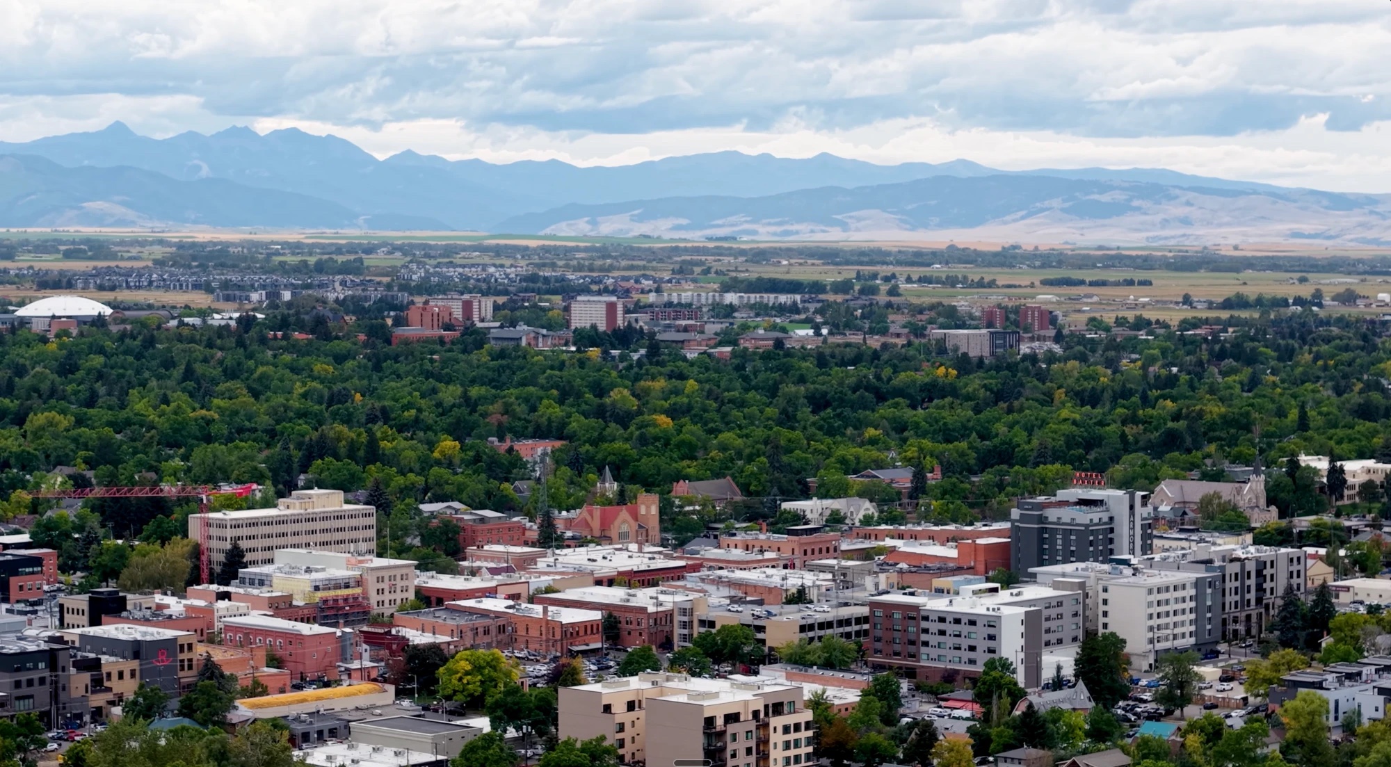 Aerial view of downtown Bozeman surrounded by trees, with the Bridger and Gallatin mountain ranges in the distance
